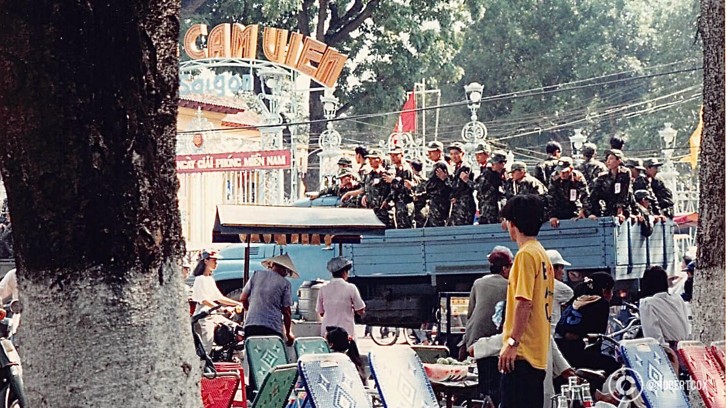 A blue truck carrying a group of soldiers, participating in the parade. The red banner with white text reads: “WELCOME THE MOVEMENT TO CELEBRATE THE YEAR.” The word “SÀIGÒN” (Saigon) is also visible on an arch, indicating the historical name of Ho Chi Minh City, often still used colloquially.(April 30, 1995)