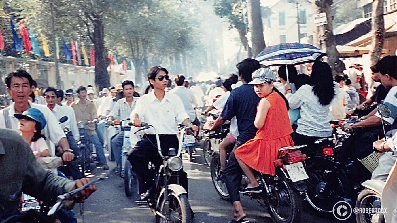 A lively scene from near the parade route in Ho Chi Minh City, Vietnam, on April 30, 1995. A large group of people on motorbikes and bicycles, moving through a tree-lined street adorned with colorful flags, near the parade procession. Some women are wearing traditional Vietnamese attire, such as the conical hat (nón lá) and bright dresses.
