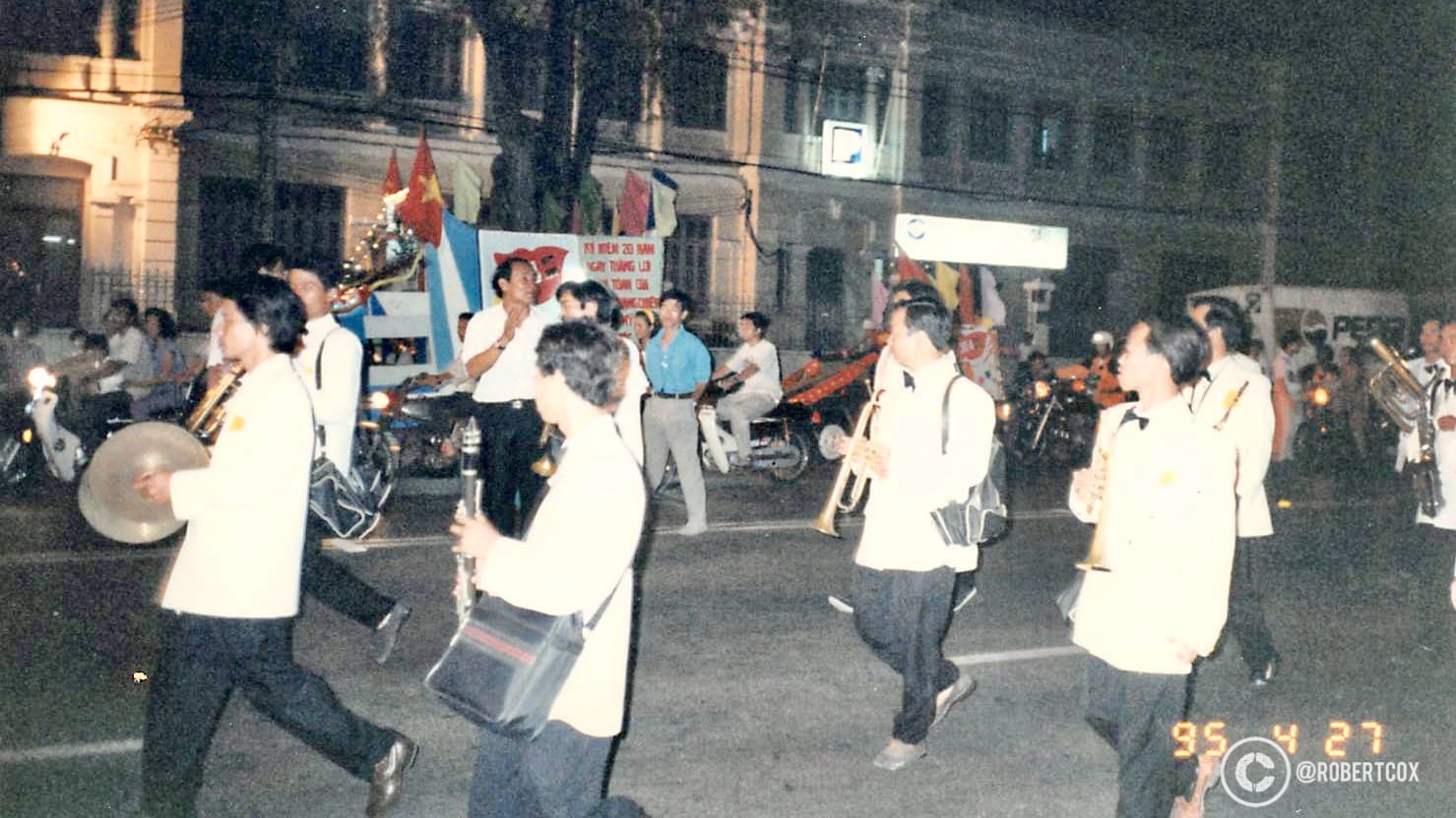 A marching band in white uniforms playing instruments like trumpets and cymbals, with Vietnamese flags and festive decorations, at the parade rehearsal on April 28, 1995.
