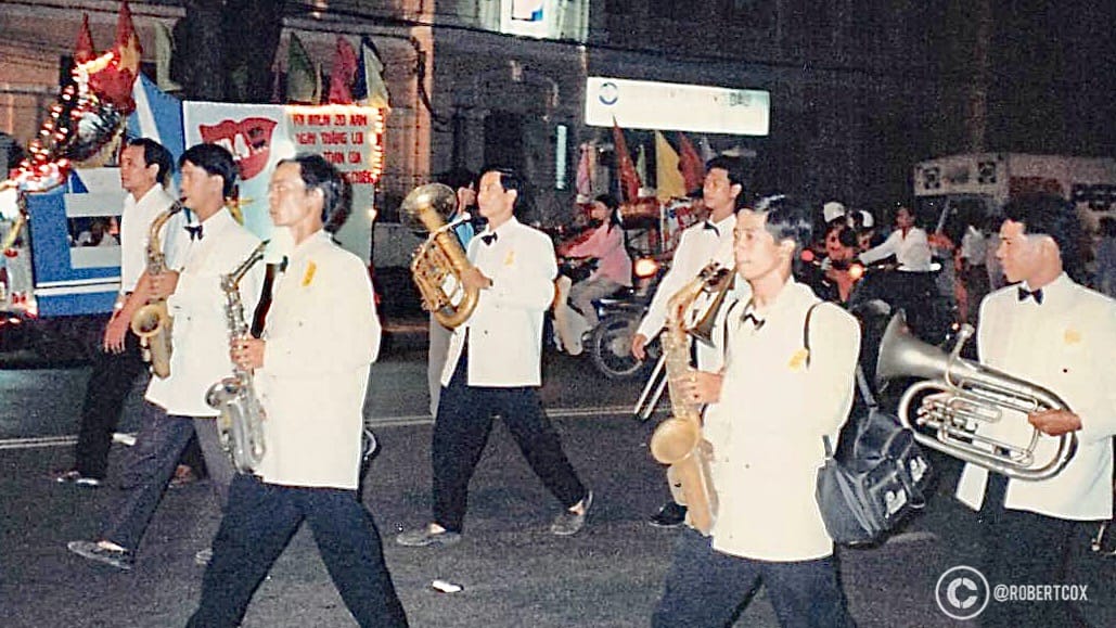 A marching band in white uniforms playing instruments like trumpets and cymbals, with Vietnamese flags and festive decorations, at the parade rehearsal on April 28,1995.
