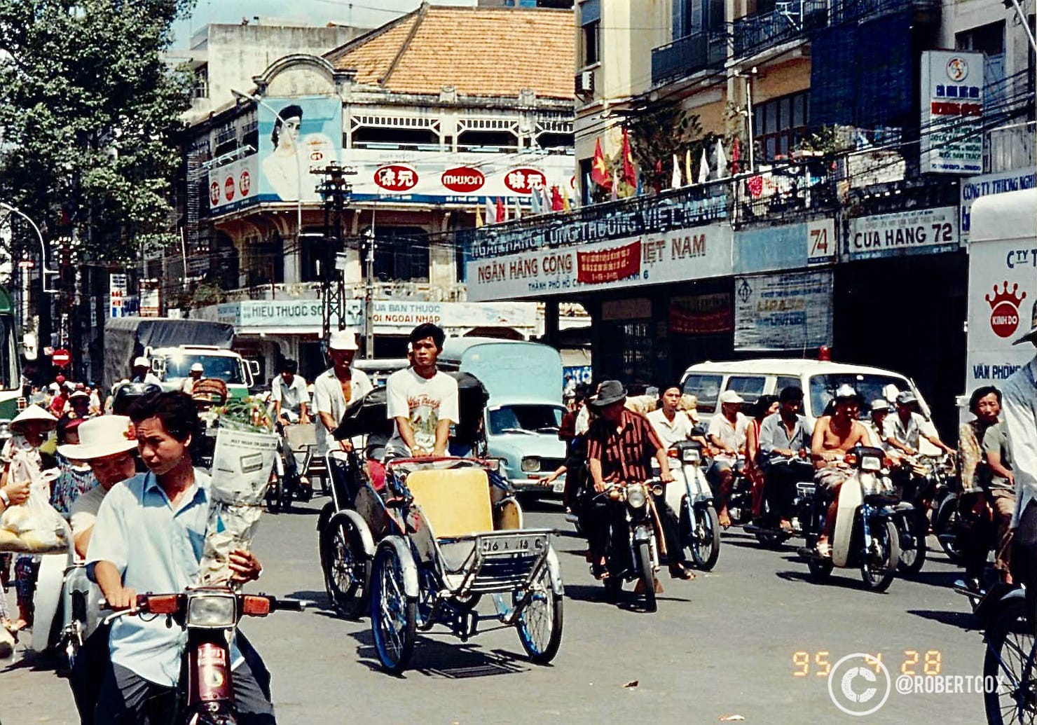 A bustling street scene in District 1 of Ho Chi Minh City (Saigon), Vietnam, during my visit for the 20th anniversary of Reunification Day. The banner translates to “Vietnam Port Bank”, a financial institution, common in central Saigon’s business districts. Motorbikes, cyclos (cycle rickshaws), and a few cars, reflect the primary modes of transportation in Saigon on April 1995, and reflects the vibrant energy of the city at the time, blending everyday life with the festive atmosphere of the celebrations.